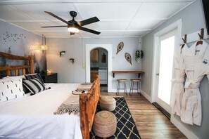 Une chambre à coucher avec un lit, une tête de lit en bois, des tables de chevet, un ventilateur de plafond, un téléviseur fixé au mur et une vue sur la cuisine.