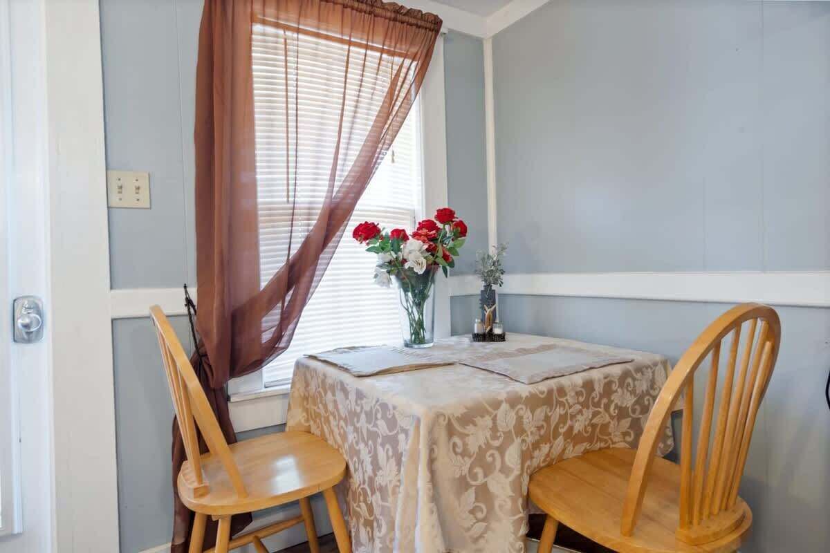 Un coin repas avec une table dressée pour deux personnes, une fenêtre avec des stores et un vase rempli de fleurs.