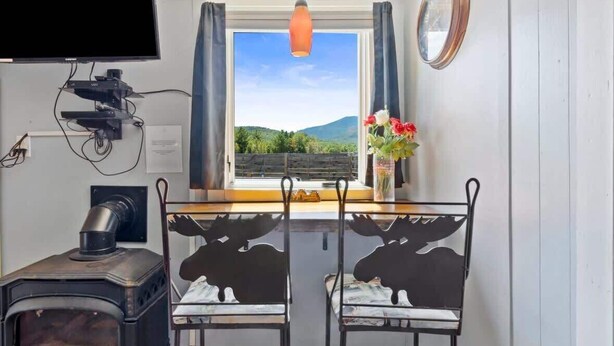 A cozy room with a wood-burning stove, a TV mounted on the wall, a dining table with two chairs, and a window with curtains.
