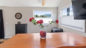 A room with a wooden table, a vase with red flowers, a clock on the wall, and a TV on the wall. - Mountain Views Hot Tub Hike Mt. Washington  #4 (Carroll)