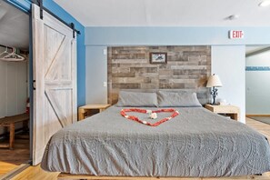 A hotel room with a bed, a wooden headboard, a heart-shaped decoration on the bed, a nightstand, a lamp, and an exit sign. - Hike White Mountain's & Relax in the Hot tub! #6 (Carroll)