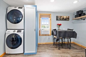 A laundry room with two washing machines, a table with chairs, a window with a view, and a shelf with bottles.