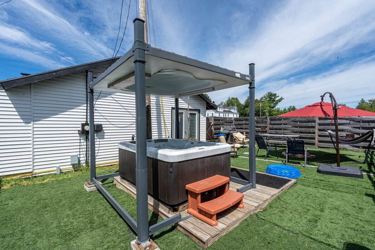 A hot tub with a wooden deck and a covered seating area, set against a house with a fenced yard.