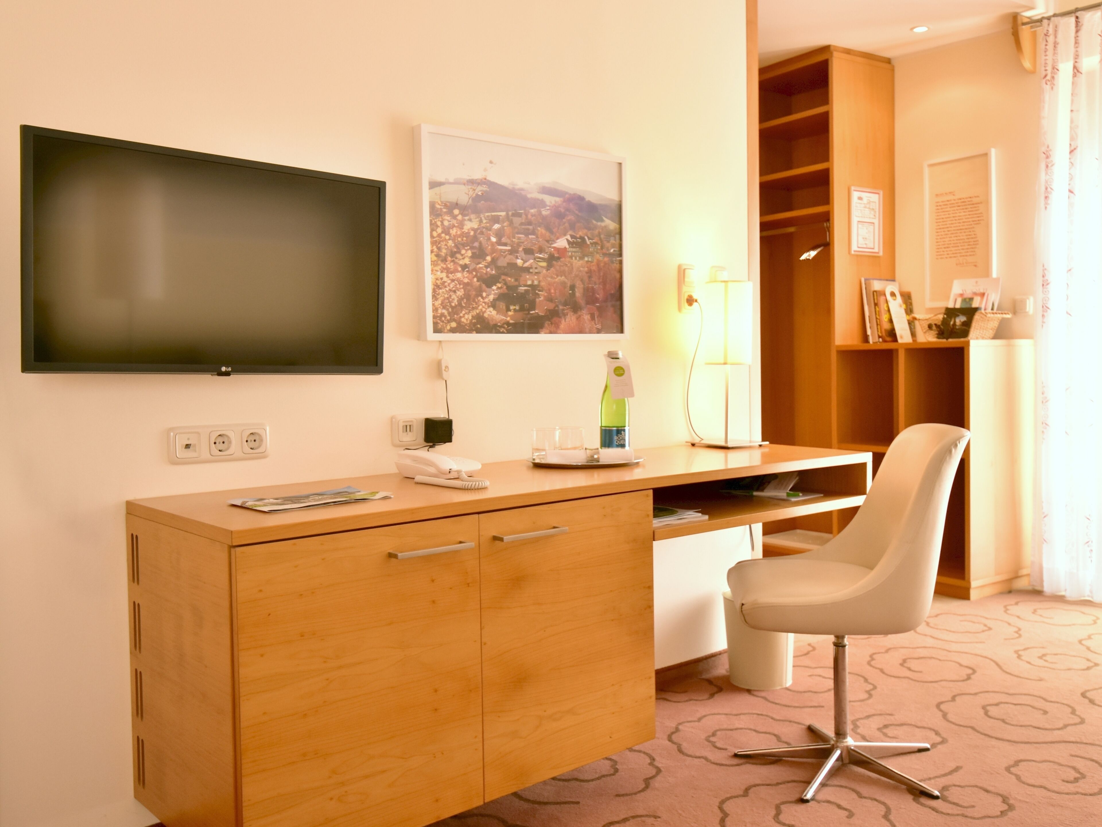 A hotel room with a wooden desk, a flat-screen TV, a cityscape painting, a lamp, and a chair.