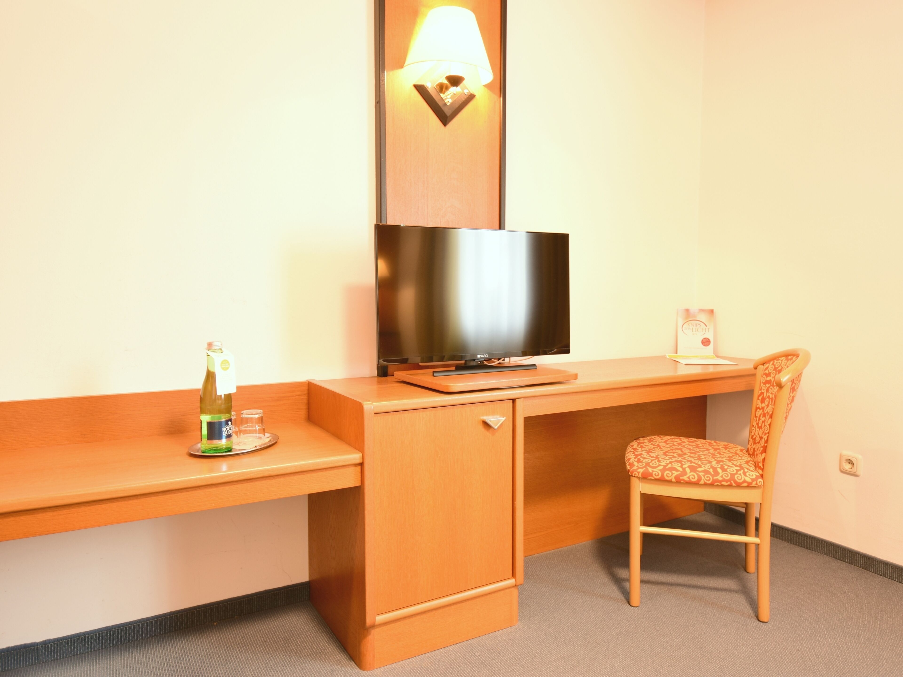 A hotel room with a wooden desk, a television, a chair, a bottle of water, and a lamp.
