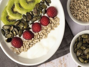 Une assiette blanche sur laquelle on voit des tranches de kiwi, des framboises et des graines de courge, le tout posé sur une surface blanche.