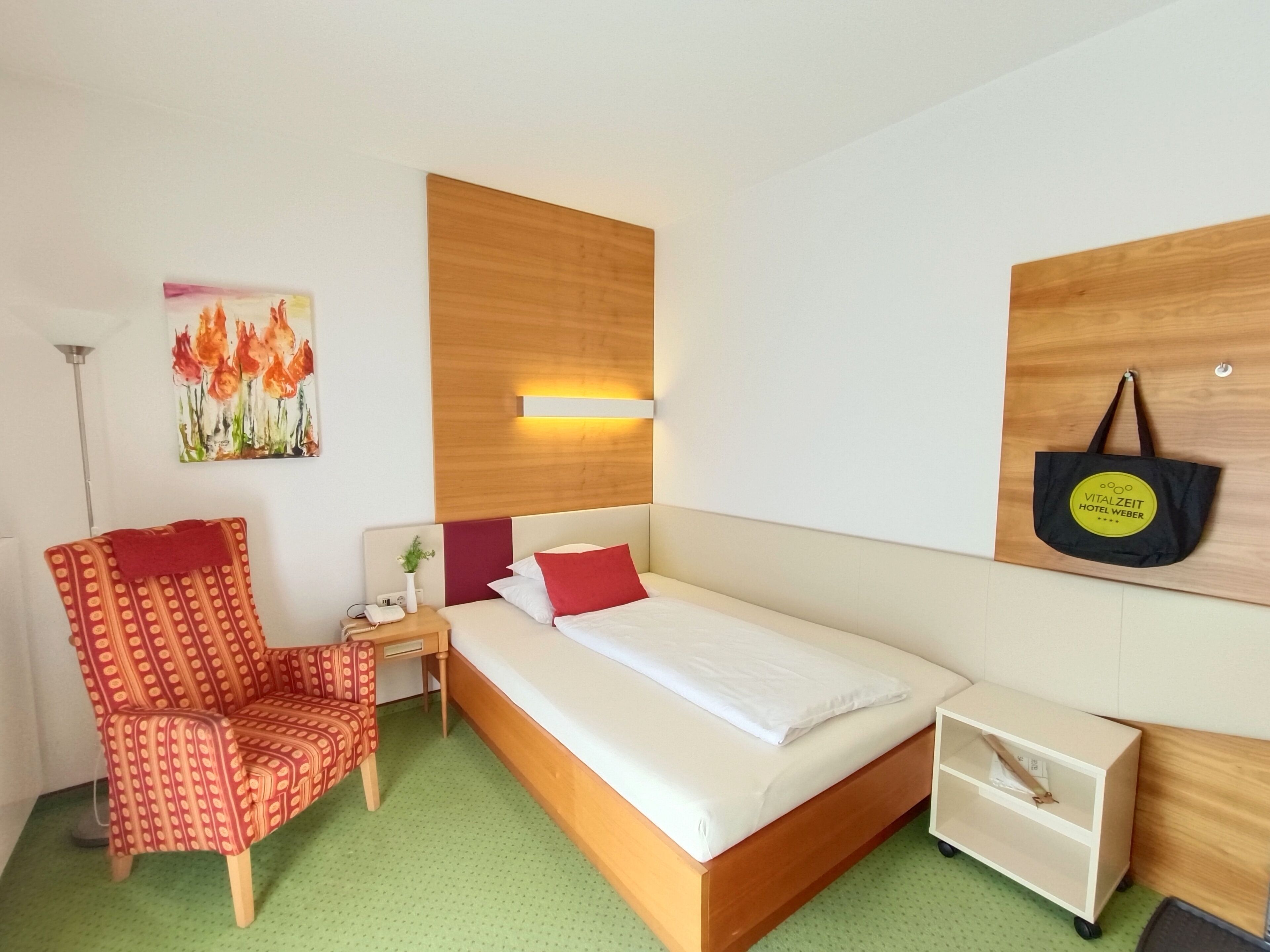 A hotel room with a bed, a chair, a nightstand, a wall-mounted lamp, and a painting.