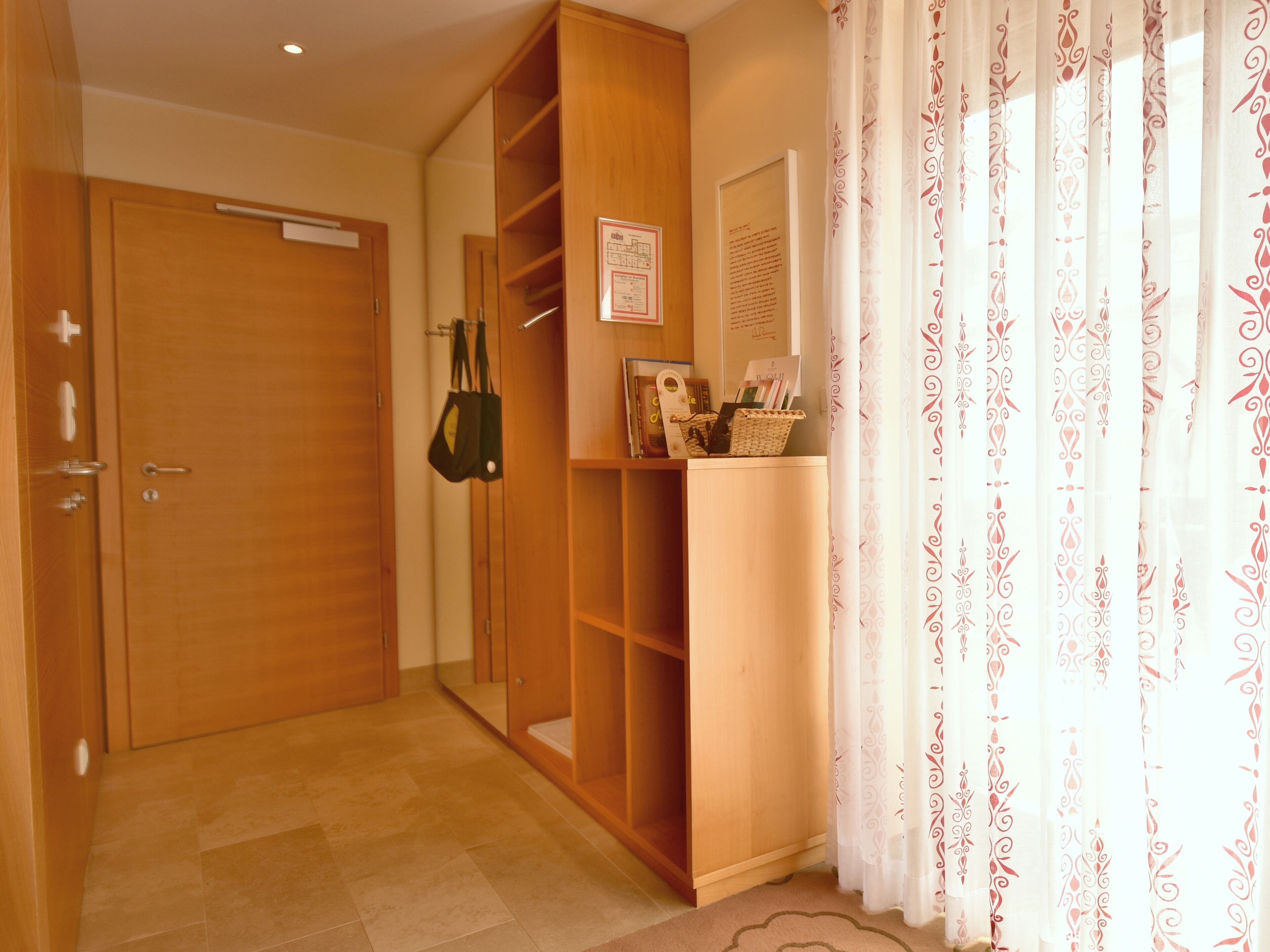 A hallway with wooden doors, a mirror, a shelf with books and a bag, and patterned curtains.