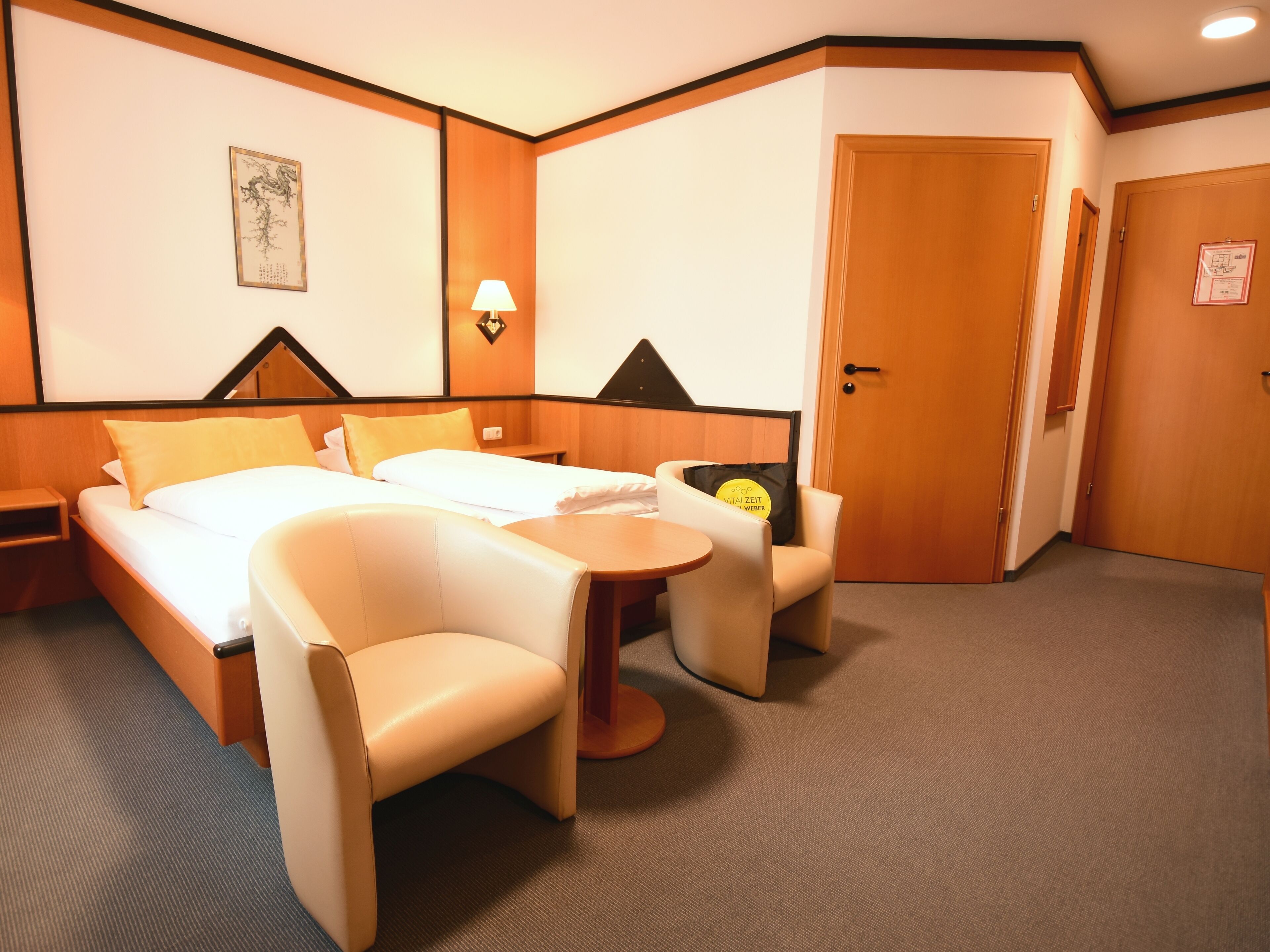 A hotel room with a bed, two chairs, a small table, a lamp, and a door.