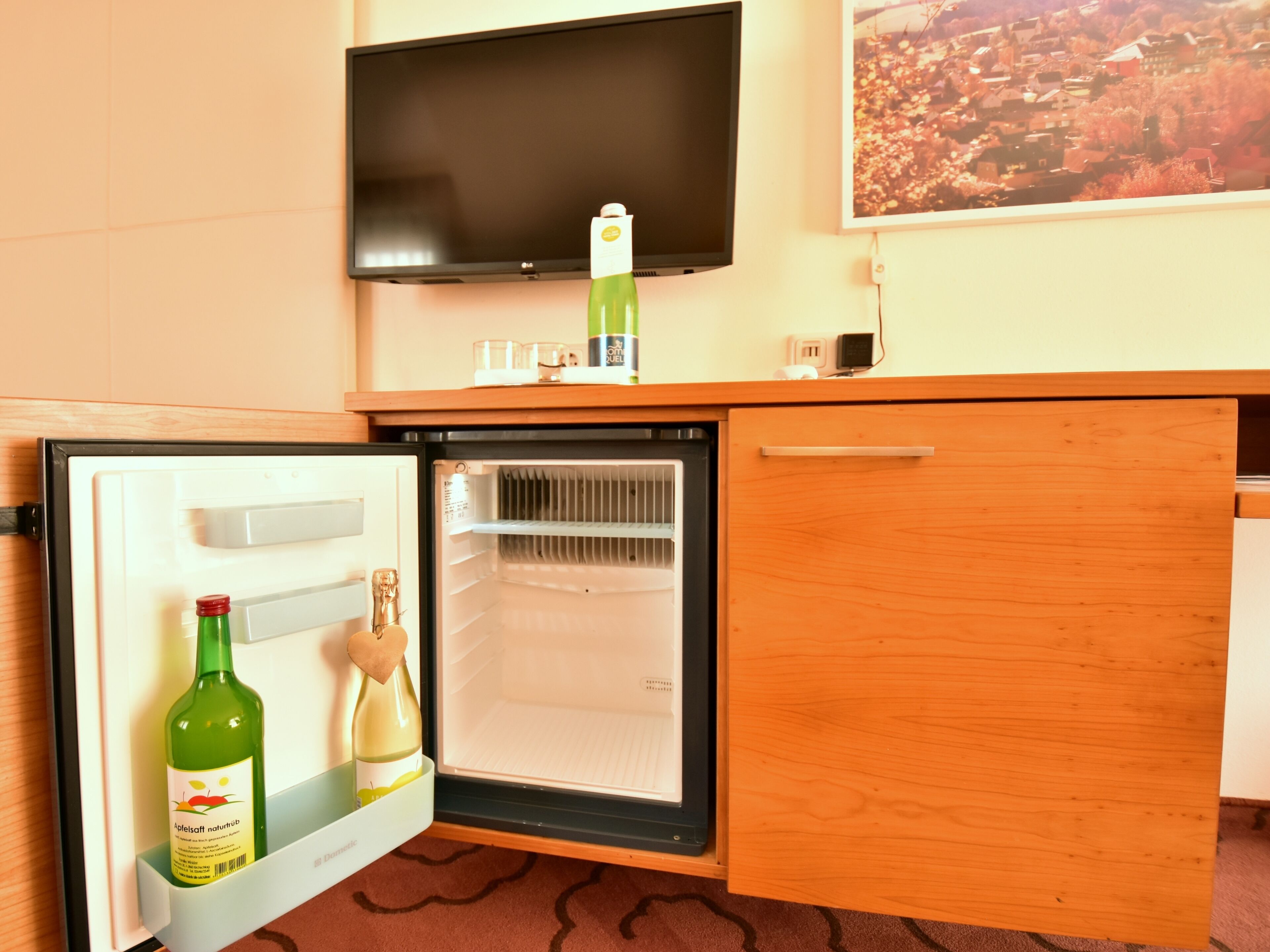 A compact kitchenette with a mini-fridge, a small refrigerator, a TV mounted on the wall, and a wooden counter.