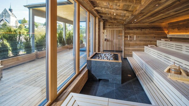 A sauna with wooden beams, stone benches, and a view of a church and greenery outside. - VitalZeit Hotel Weber (Bad Schönau)