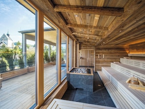 Una sauna con vigas de madera, bancos de piedra y vistas a una iglesia y follaje exterior.