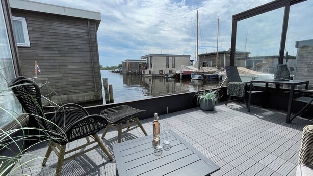 Una terraza con mobiliario exterior, una planta en maceta y vistas a barcos y edificios al otro lado del agua.