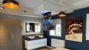 Un dormitorio moderno con una cama, un tocador con dos lavamanos, un espejo grande, un televisor de pantalla plana y un cuadro en la pared.