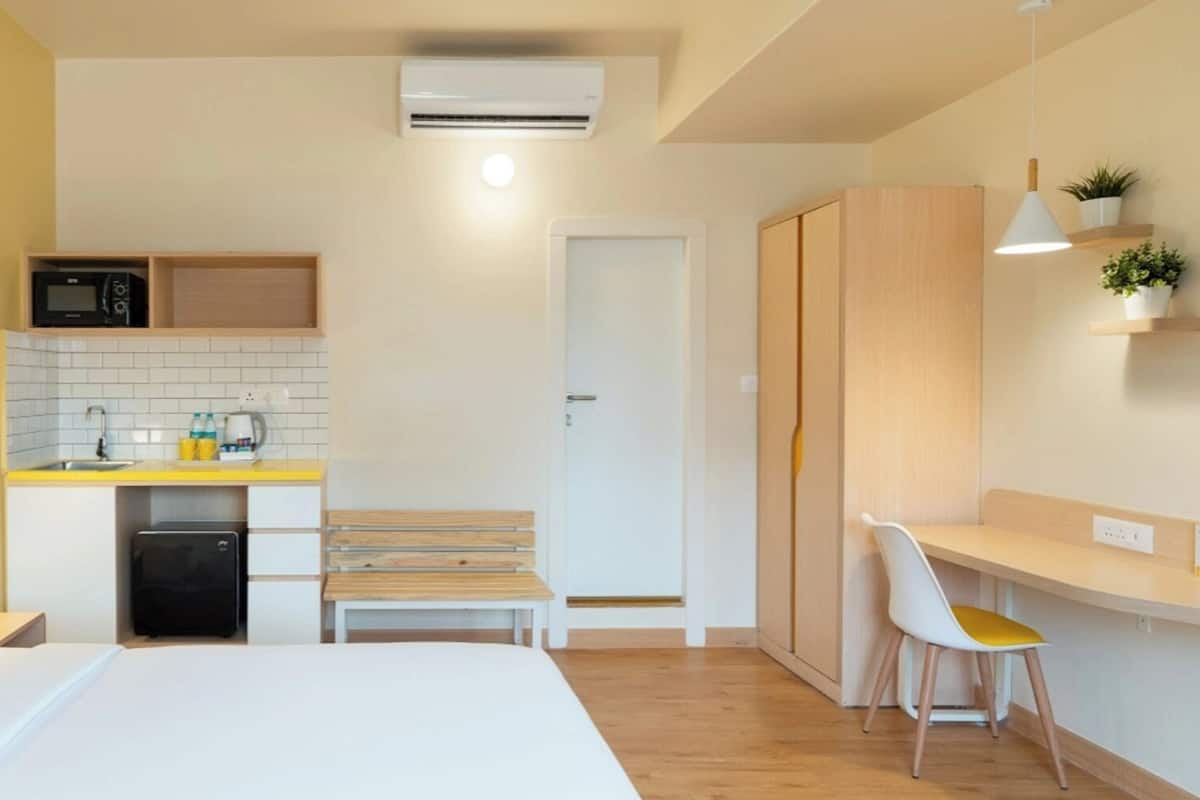 Ett kompakt hotellrum med en säng, ett sängbord, ett litet pentry, ett skrivbord med en stol och en garderob.