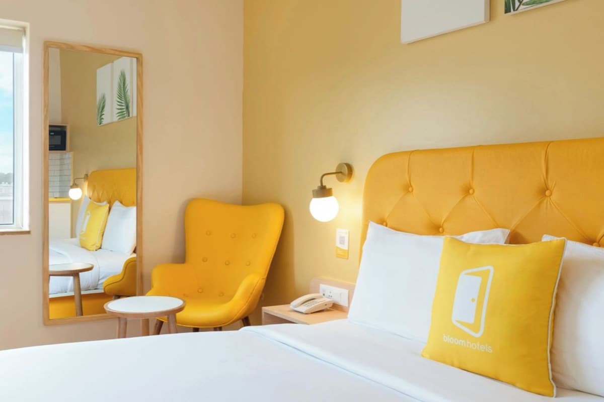 A hotel room with a bed, a chair, a small table, a mirror, and a window with a view of buildings.