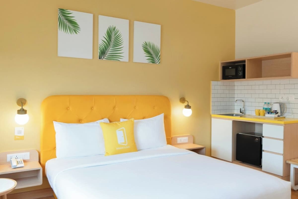 A hotel room with a bed, a yellow armchair, a small desk, a nightstand, a wall-mounted TV, and a kitchenette.