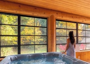 A room with a large window, a hot tub, and a person standing by the window.
