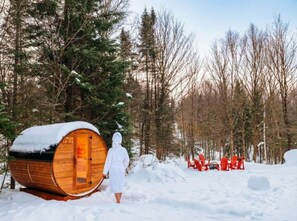 Una cabaña de madera con exterior cubierto de nieve, una persona vestida de blanco y sillas rojas al fondo.