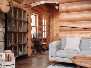 A log cabin interior with a sofa, a wooden cabinet with glass doors, a chair, and a hanging plant. - Serenity / SPA / Sauna / Fiddler Lake (Mille-Isles)