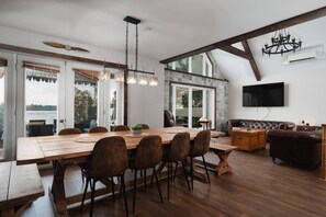 Uma sala de jantar espaçosa com uma grande mesa de madeira e cadeiras, um candelabro e vista para o exterior através de amplas janelas.