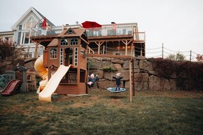 A wooden playhouse with a slide and a swing set in a backyard.