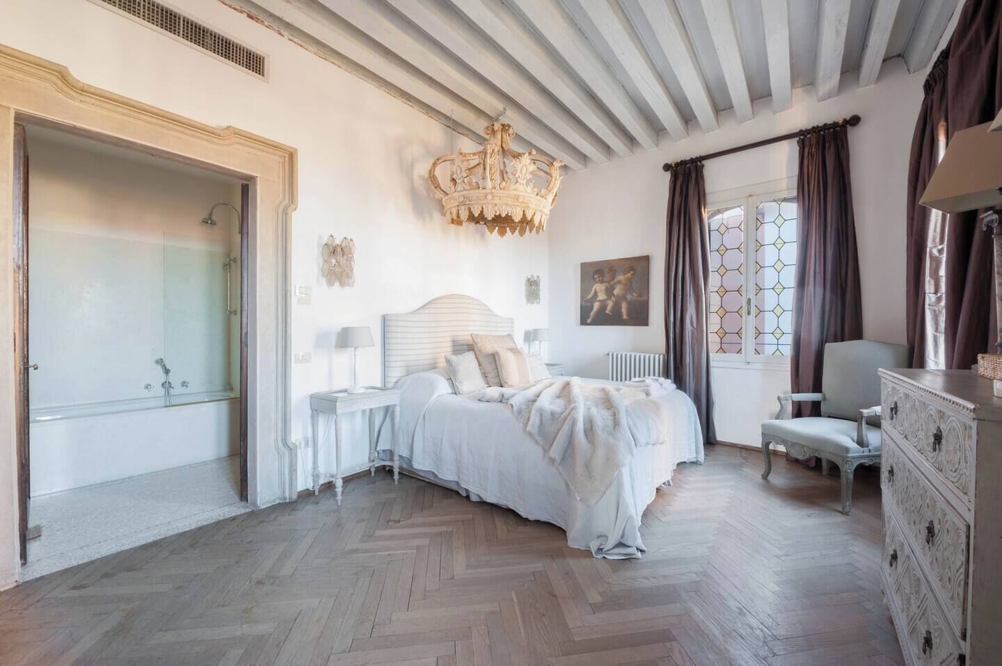 A bedroom with a large bed, a chandelier, a dresser, and a window with curtains.