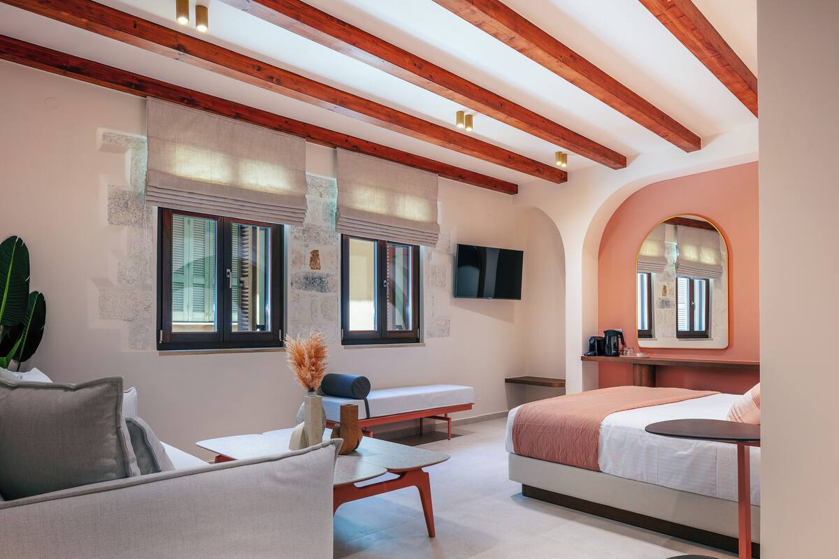 A modern hotel room with a bed, a sofa, a small table, and a bench. There is a TV mounted on the wall and a window with a view of the outside.