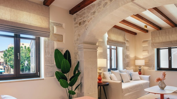 A room with a stone archway, a white sofa, a small table with a vase, and a bed with a pink quilt. - La Montre Luxury Suites (Rethymno)