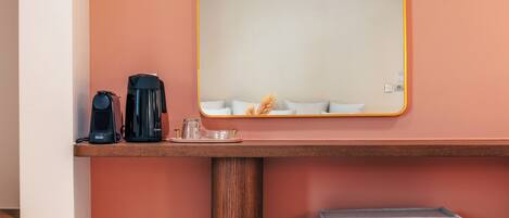 A hotel room with a wooden bedside table, a black coffee maker, a glass pitcher, and a mirror with a yellow frame.