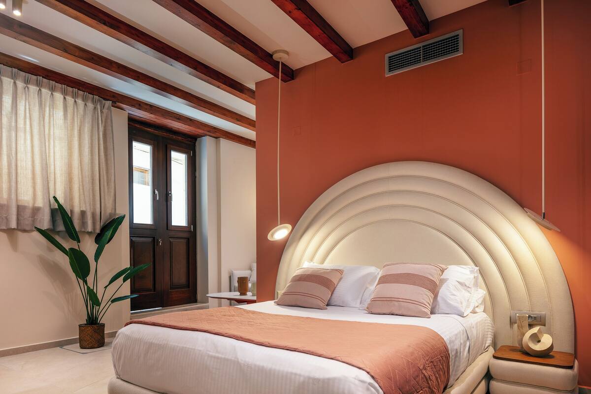 A bedroom with a large bed, a bedside table, a potted plant, and a window with curtains.