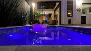 Una piscina con una luz flotante, rodeada de plantas y un edificio al fondo.