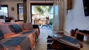 Un salón acogedor con un sofá, una mesa de centro con libros, una mesita con una cesta y vistas a una piscina y palmeras afuera.