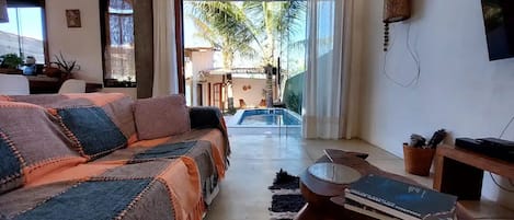 A cozy living room with a sofa, a coffee table with books, a small table with a basket, and a view of a pool and palm trees outside.