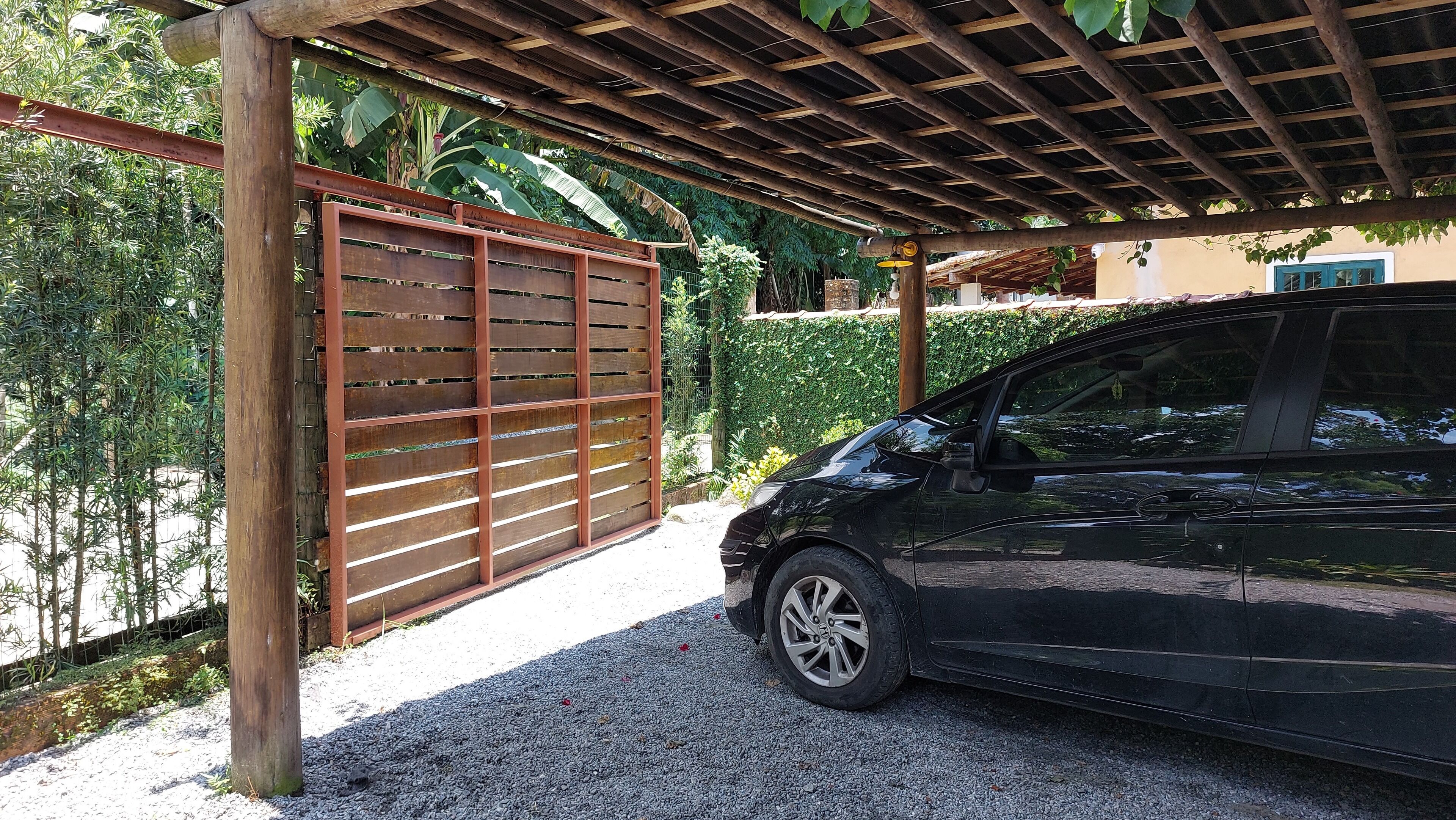 Een zwarte auto geparkeerd onder een houten pergola, met een metalen poort op de achtergrond.
