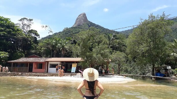 Una playa con una montaña al fondo, un edificio de techo rojo y gente en el agua.