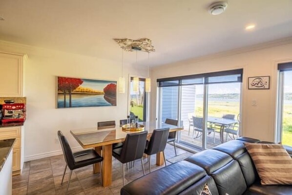 A dining area with a table and chairs, a painting on the wall, and a view through large windows. - Waterfront view | Phenicia House (Sainte-Anne-de-Beaupré)