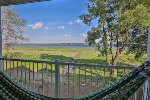 A hammock on a balcony overlooking a grassy landscape with trees and a body of water.