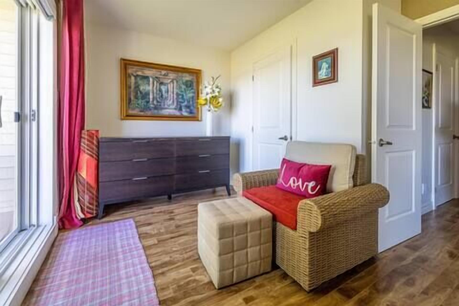 Una habitación acogedora con un sofá de mimbre, un pequeño taburete, un tapete de cuadros, un aparador oscuro y un cuadro en la pared.