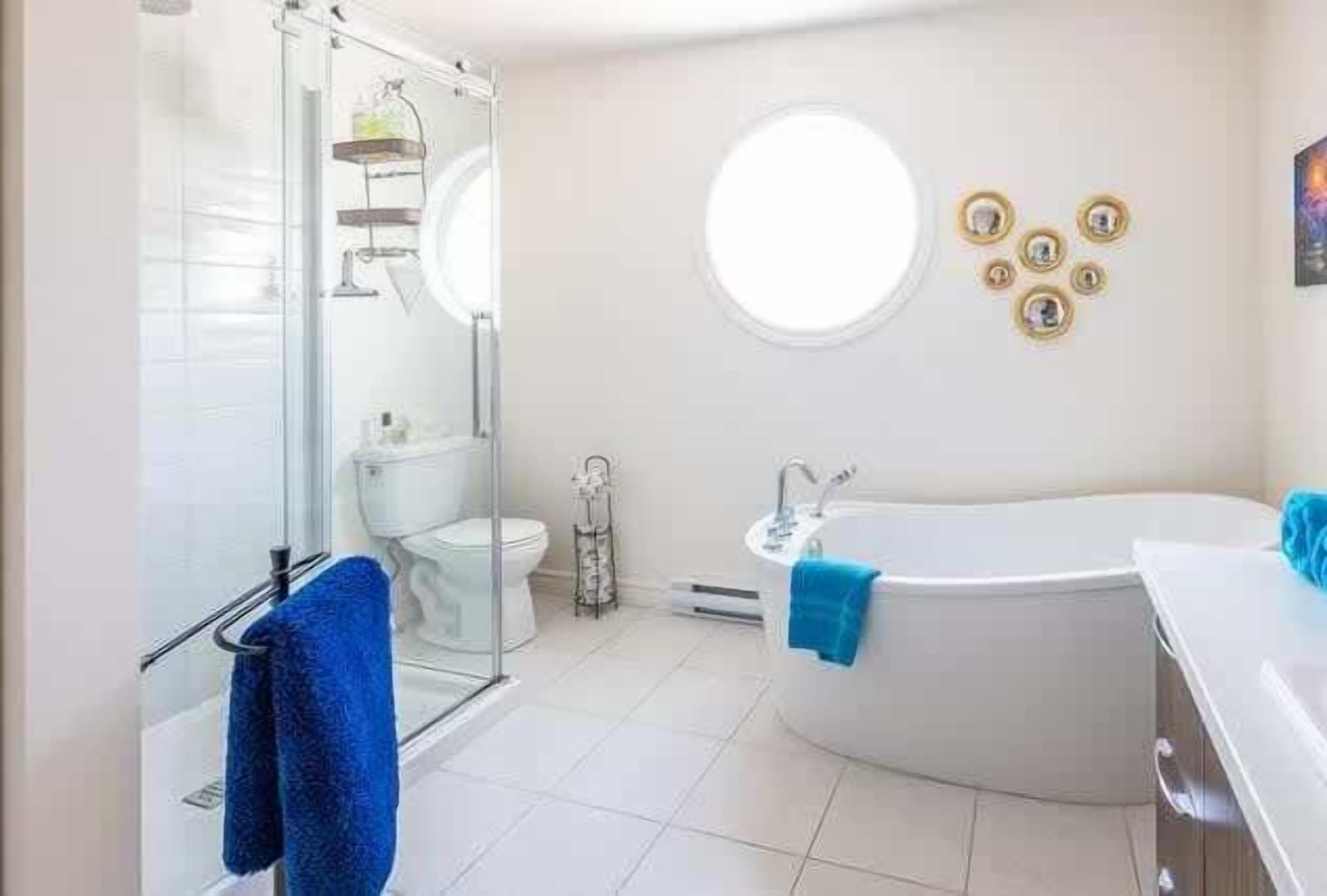 A modern bathroom with a glass shower enclosure, a white toilet, a round mirror, a bathtub, and a small round window.