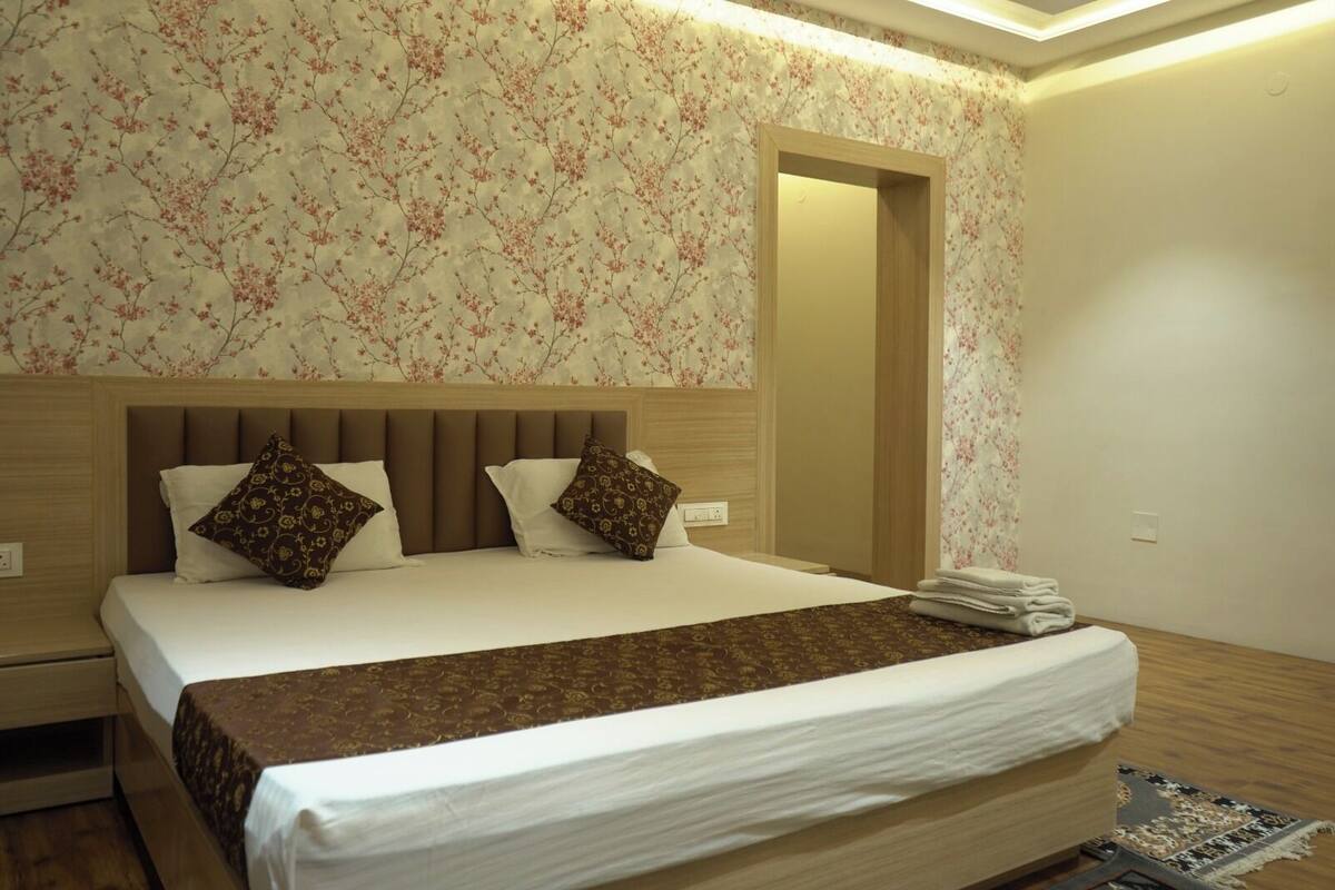 Una cama bien hecha con una colcha marrón y dos almohadones estampados, un cabecero de madera y papel tapiz con motivos florales.