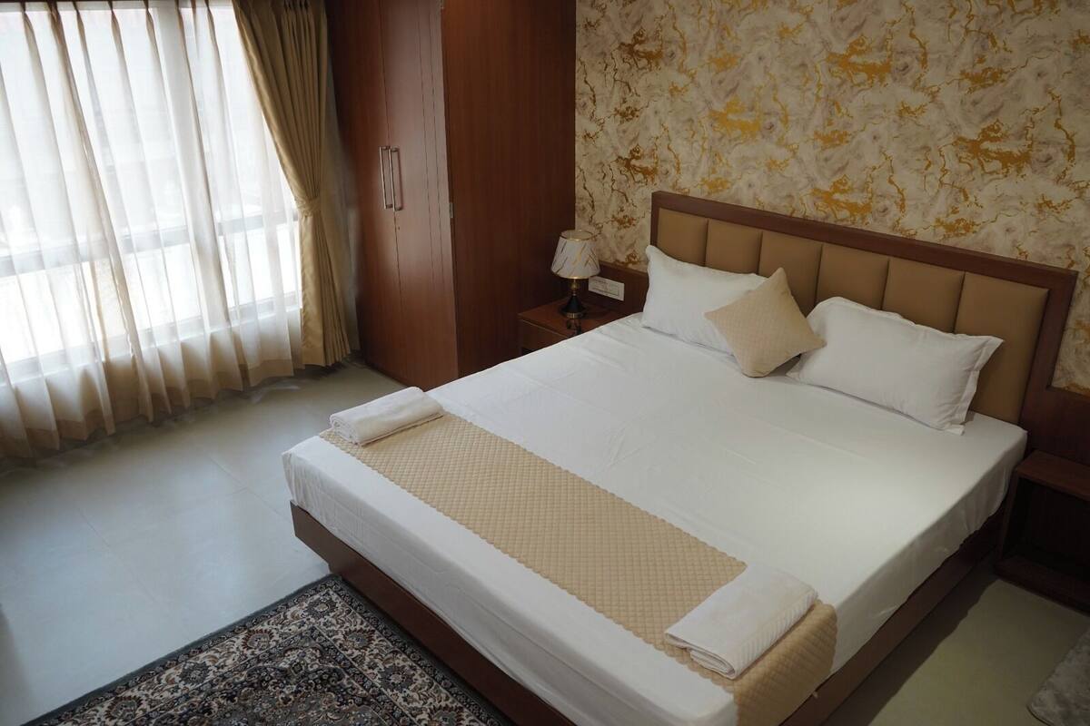 Una cama bien hecha con colcha acolchada y almohadas, una mesita de noche con lámpara, un armario de madera y una alfombra estampada.