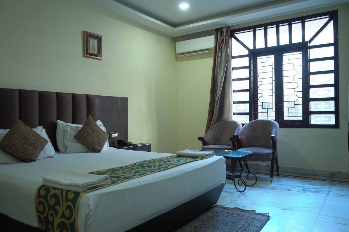 Una habitación de hotel con una cama grande, dos sillones, una mesita, un ventanal con vistas y un equipo de aire acondicionado.