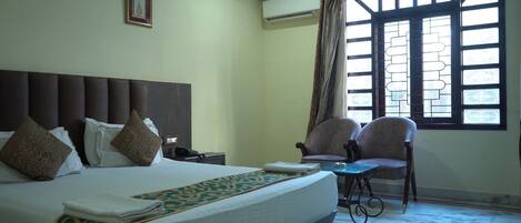 A hotel room with a large bed, two armchairs, a small table, a window with a view, and an air conditioning unit.
