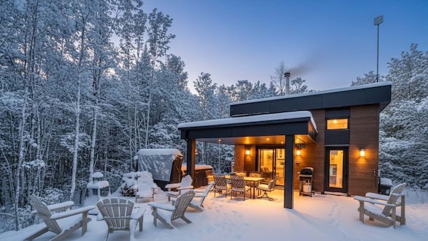 Una cabaña moderna con un espacio de estar al aire libre cubierto, rodeada por un bosque nevado.