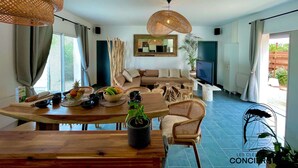 A spacious living area with a wooden dining table, wicker chairs, and a large woven light fixture. A comfortable sofa and a flat-screen TV are also visible.