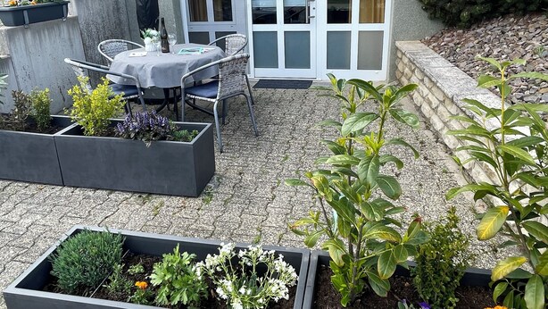 Une terrasse extérieure avec une table et des chaises, entourée de jardinières surélevées où sont plantées différentes espèces.