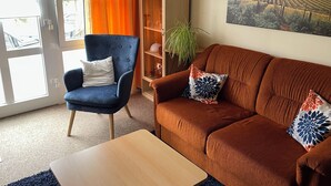 Un salon avec un fauteuil bleu, un canapé marron orné de coussins à motifs, une table basse en bois et une bibliothèque décorée de divers objets.
