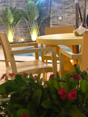 An outdoor seating area with wooden chairs and a table, surrounded by large green plants and a brick wall. - Pousada Velosmar (Ipojuca)