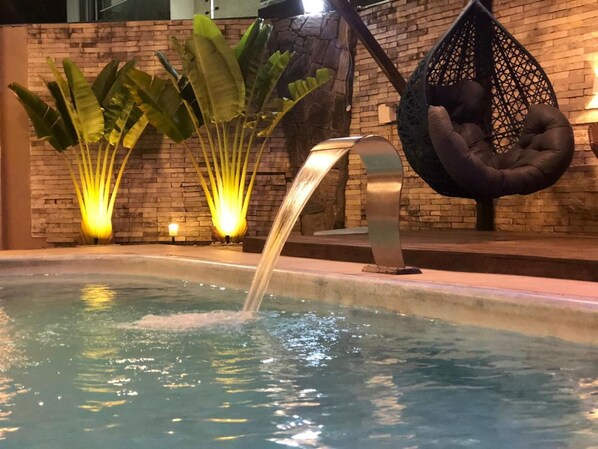 A pool with a water feature, surrounded by plants and a hanging chair. - Pousada Velosmar (Ipojuca)